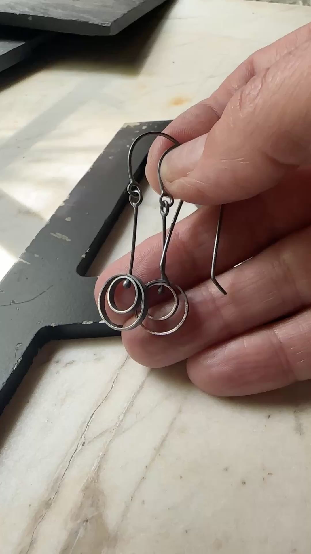 A pair of oxidized sterling silver drop earrings with circular spiral designs, resting on a light beige marble surface next to a black wooden frame.