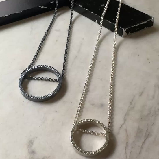 One large oxidized hammered silver circular pendant on a dark silver chain and one bright silver hammered silver circle pendant on a bright silver chain