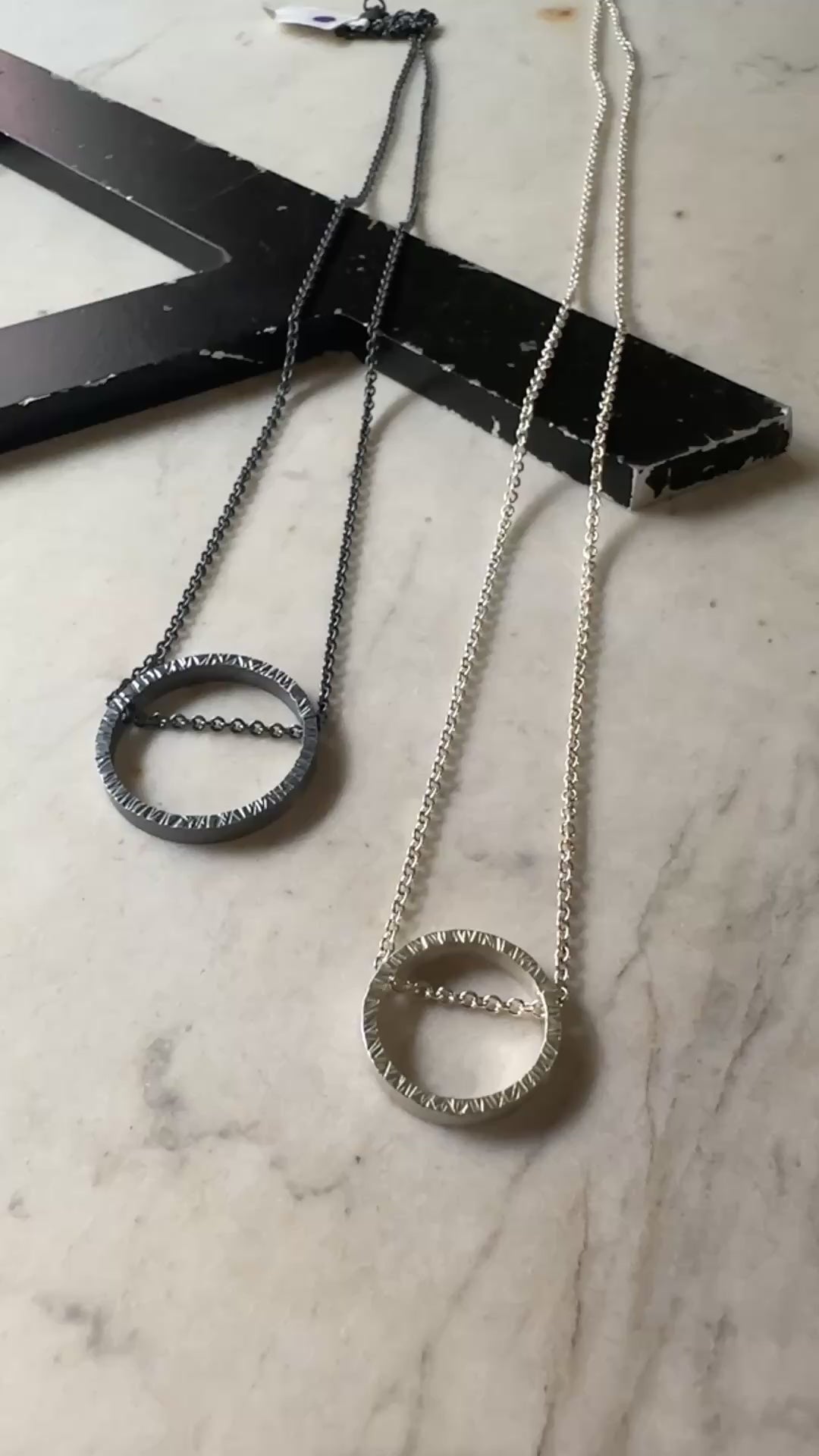 One large oxidized hammered silver circular pendant on a dark silver chain and one bright silver hammered silver circle pendant on a bright silver chain