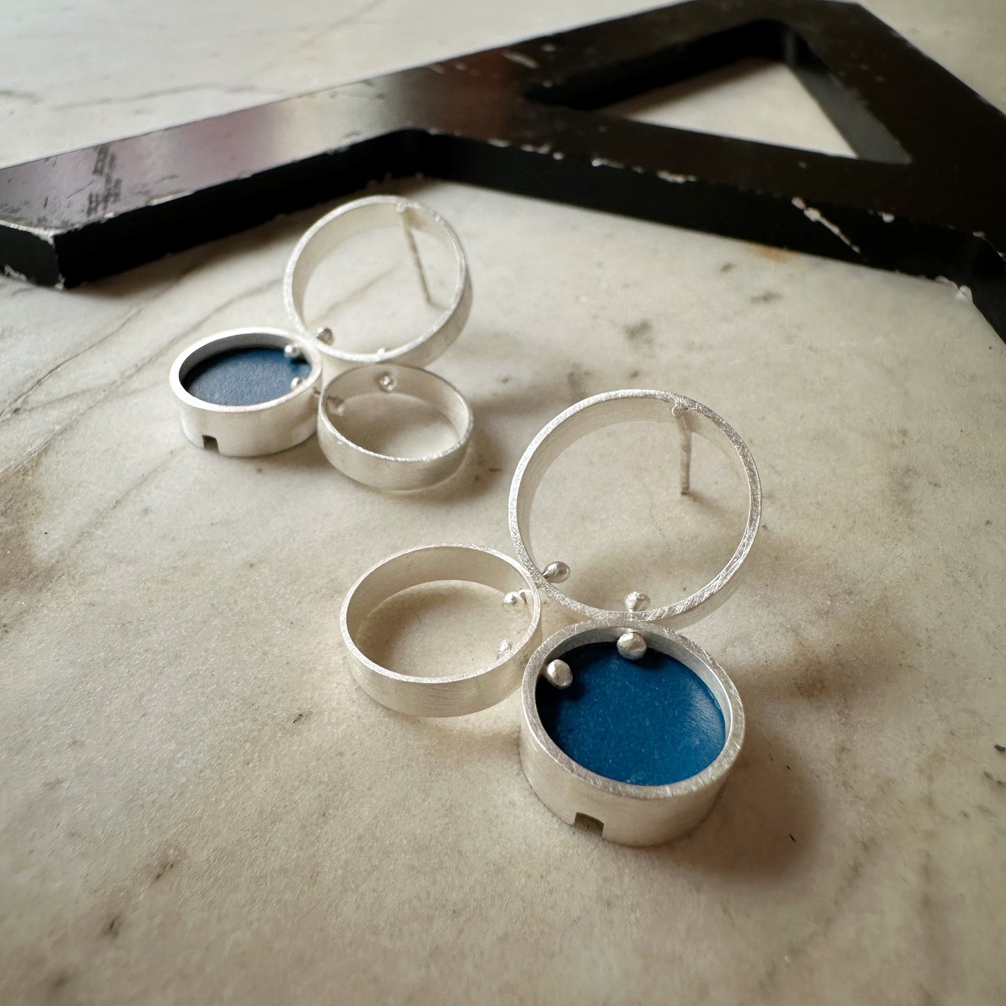 A pair of silver post  earrings with cellular drop elements featuring small round blue formica, displayed on a light marble surface.