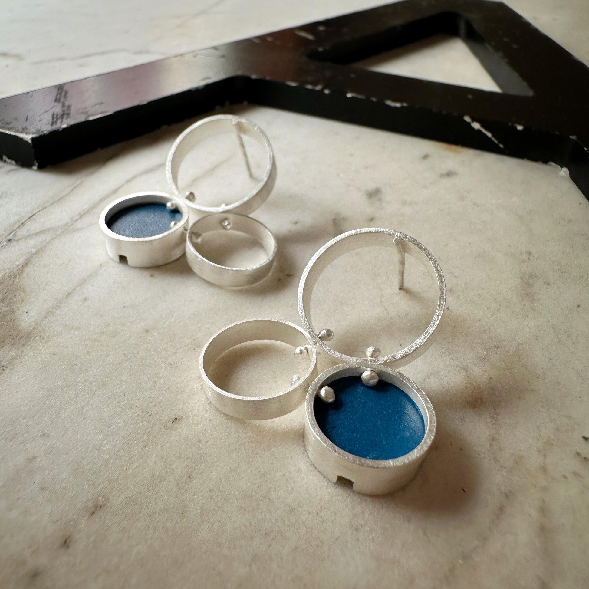 A pair of silver post  earrings with cellular drop elements featuring small round blue formica, displayed on a light marble surface.
