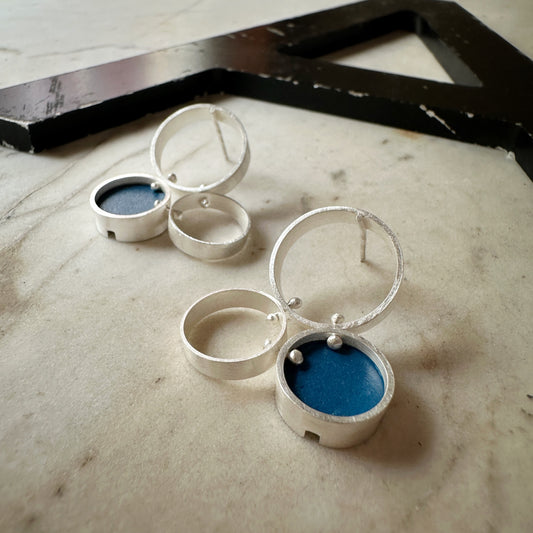 A pair of silver post  earrings with cellular drop elements featuring small round blue formica, displayed on a light marble surface.