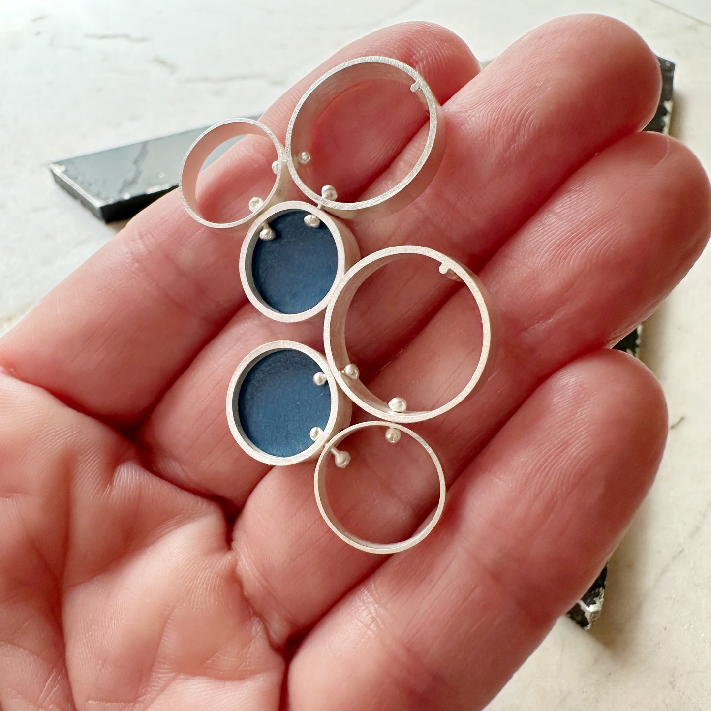 A pair of silver post  earrings with cellular drop elements featuring small round blue formica, displayed on a hand