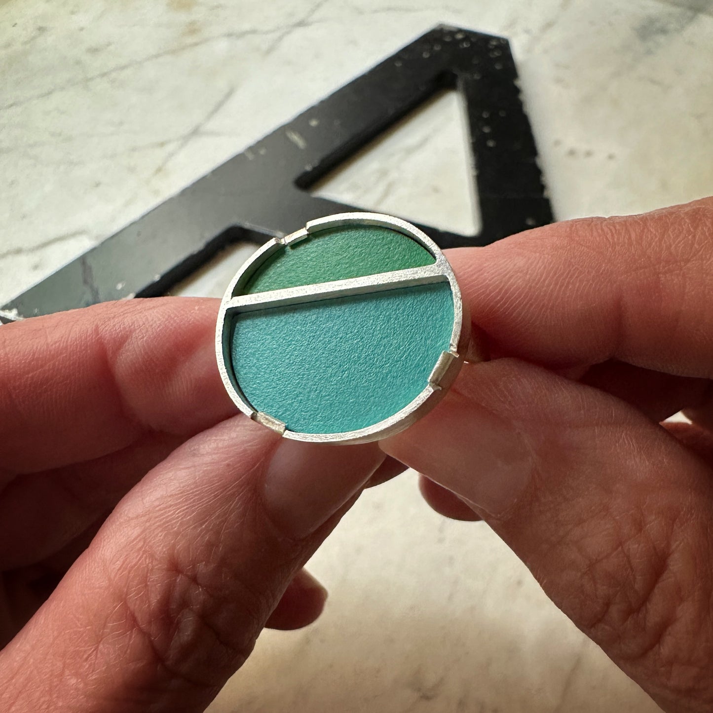 A close-up of a hand holding a circular silver ring with a two-tone green formica inlay, showing one half in a lighter mint green and the other in a darker forest green.