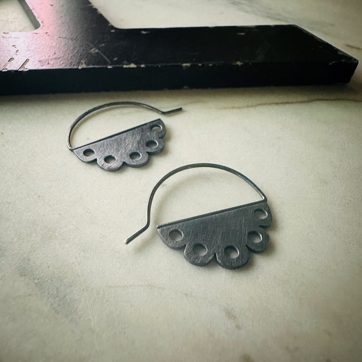 one pair of oxidized silver earrings with a scalloped edge and a pierced design on a marble surface