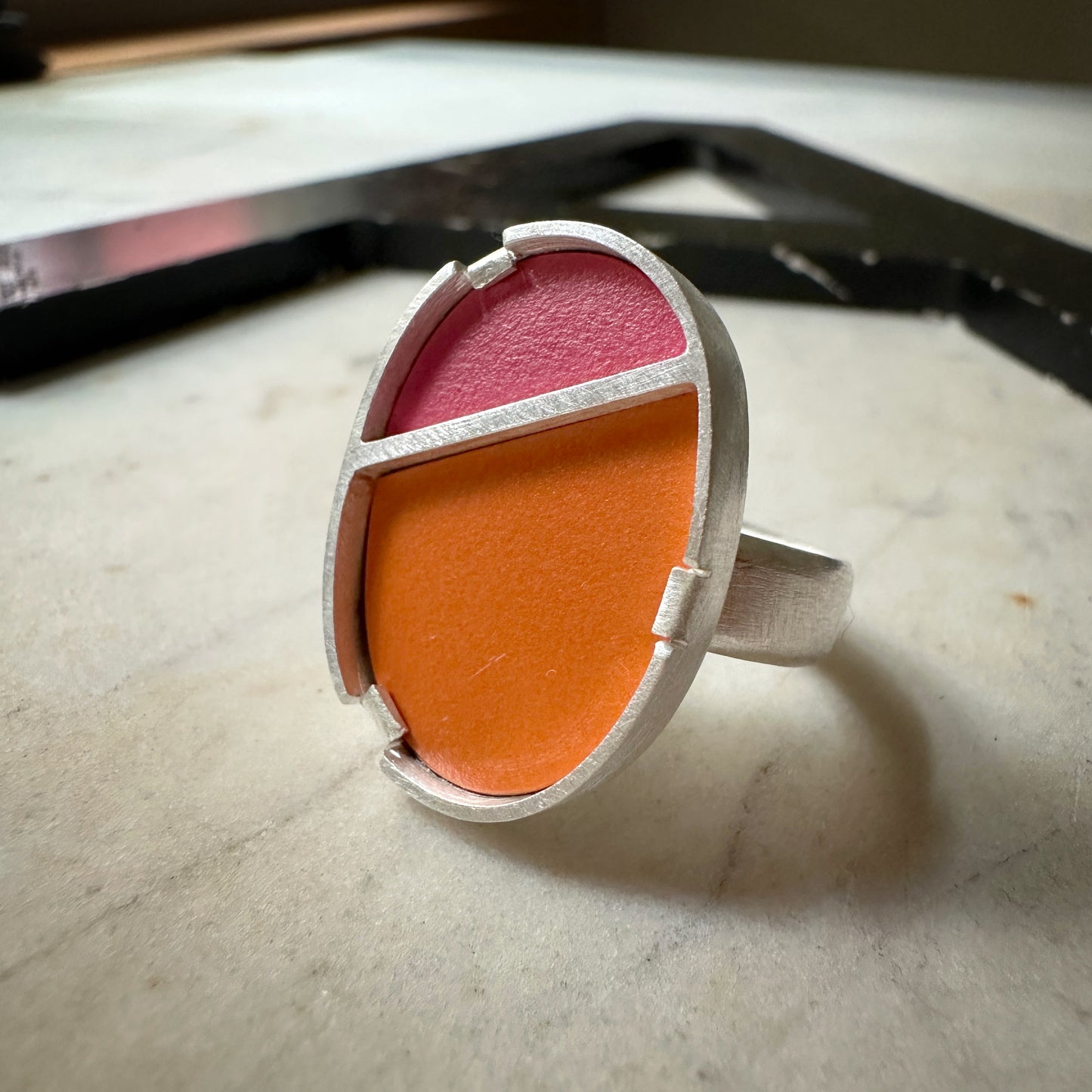 A silver ring with a geometric, segmented oval face featuring a bright orange lower half and a pink upper half, resting on a light marble surface.
