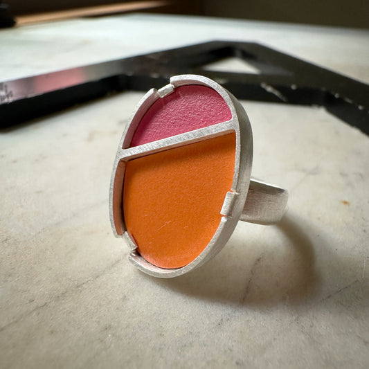 A silver ring with a geometric, segmented oval face featuring a bright orange lower half and a pink upper half, resting on a light marble surface.
