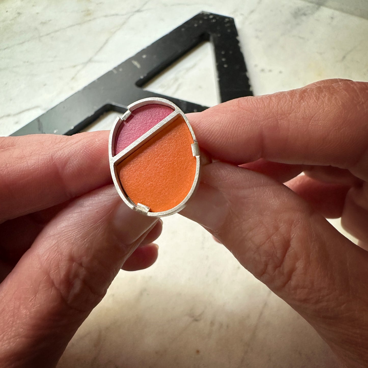 A close-up of a person's hands holding a silver oval ring with two compartments of pressed powder makeup, one pink and one orange, against a light marble background.