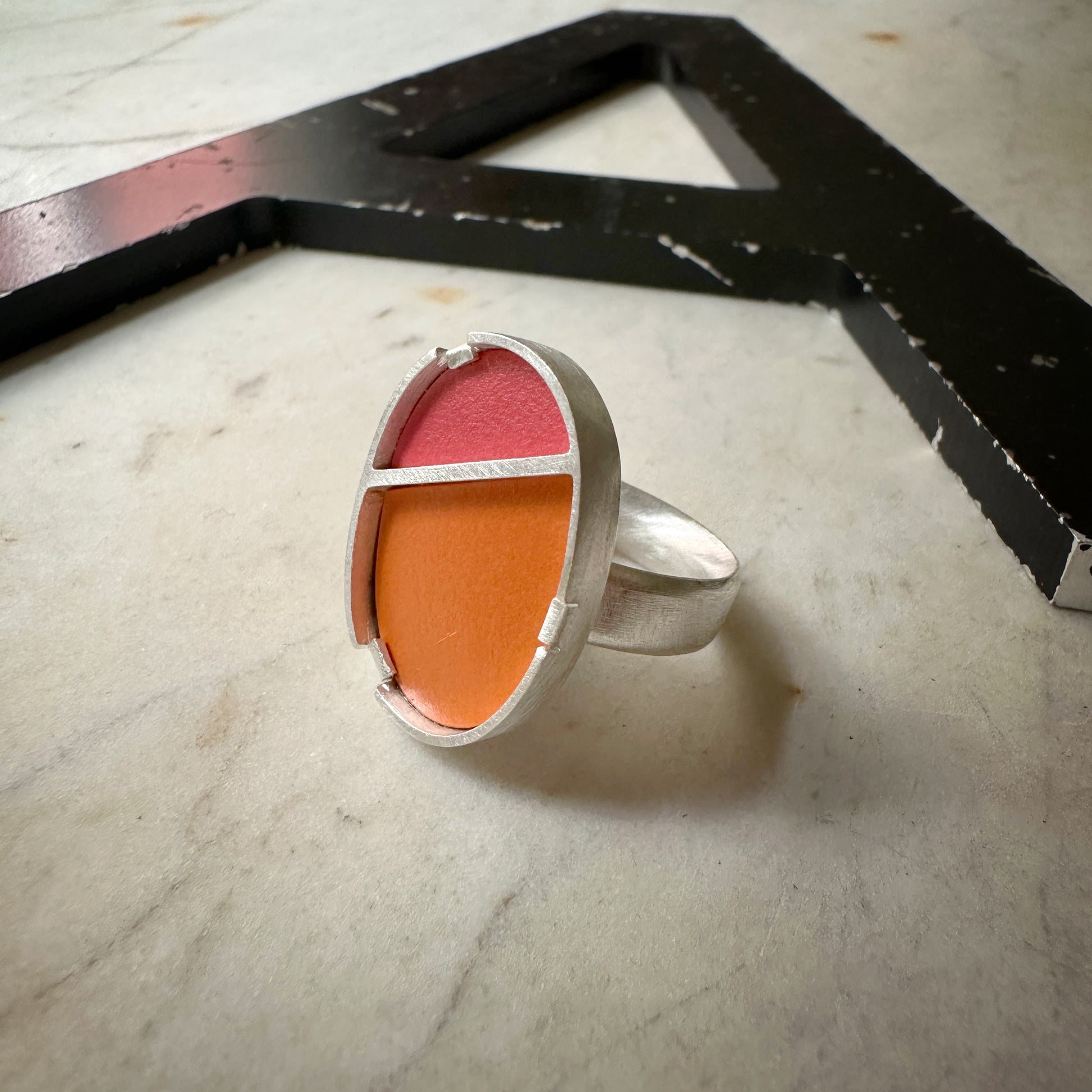 A silver ring with a large, rounded rectangular face divided into two color sections: a pink top and an orange bottom. The ring is photographed on a light marble surface next to a black metal letter 'A'.