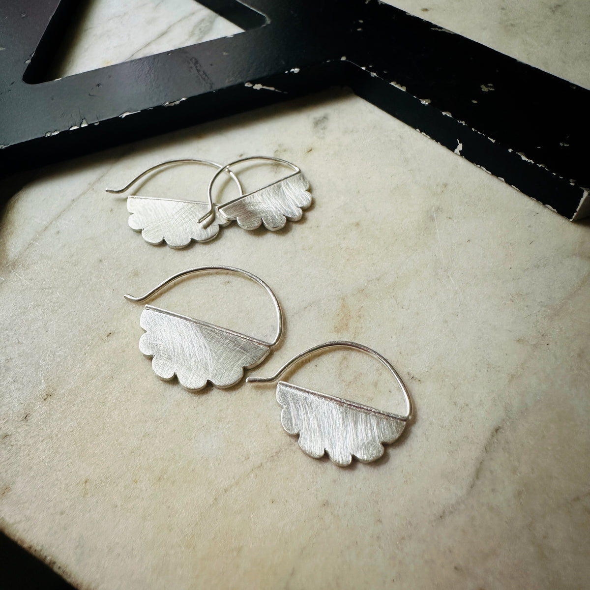 two sizes of  silver cloud-shaped hoop earrings with a textured finish, arranged on a light beige marble surface next to a black wooden frame.
