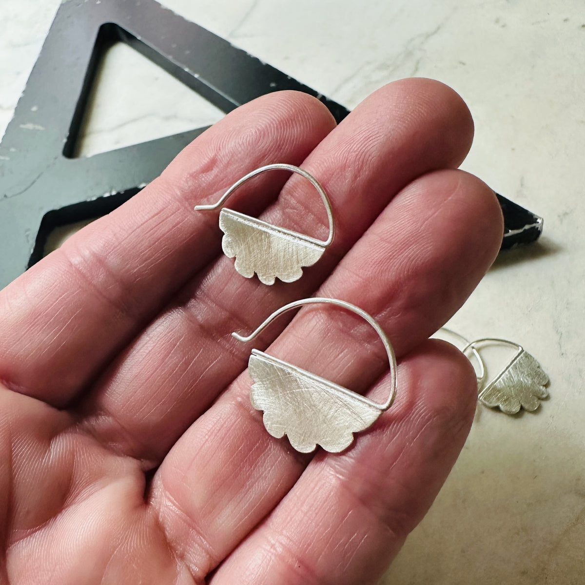 A pair of textured silver cloud shaped hoop earrings held in a person's hand, with a metal square tool visible in the background on a light stone surface.