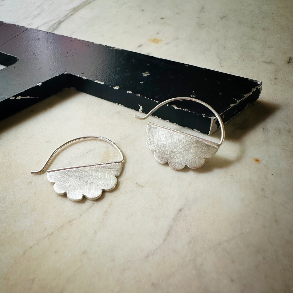 A pair of silver cloud shaped hoop earrings with scalloped edges, displayed on a light beige marble surface next to a black wooden block.