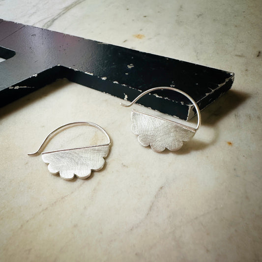 A pair of silver cloud shaped hoop earrings with scalloped edges, displayed on a light beige marble surface next to a black wooden block.