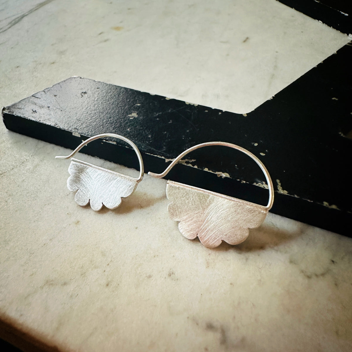 A pair of silver hoop earrings with textured, scalloped cloud-shaped dangles, displayed on a black wooden block over a light marble surface.