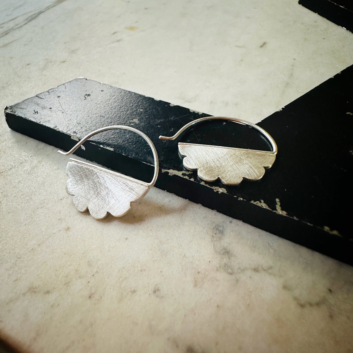 A pair of silver hoop earrings with textured, cloud-like scalloped halves, displayed on a black wooden block over a light beige marble surface.