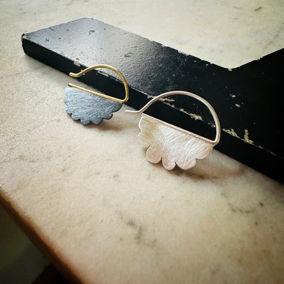 A pair of sculptural, asymmetrical hoop earrings with textured cloud-shaped drops, one in brushed gold and one in brushed silver, displayed on a black wooden block over a beige marble surface.