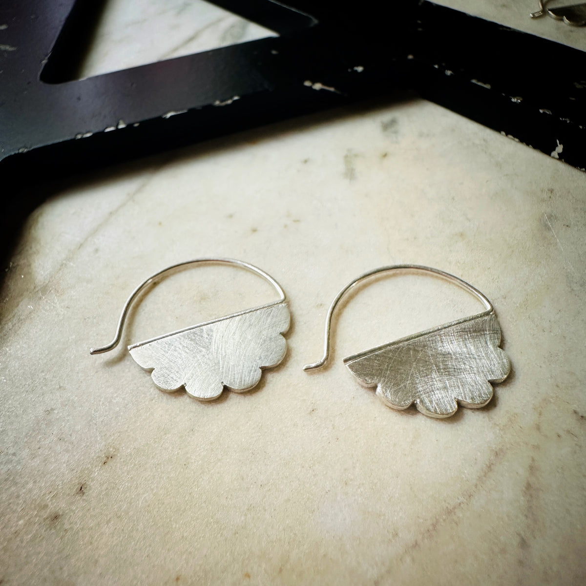 A pair of textured sterling silver cloud shaped hoop earrings with scalloped lower edges, displayed on a light beige stone surface.