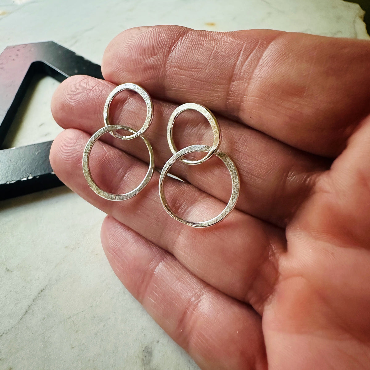 A pair of textured silver hoop earrings held in a person's hand, shown against a light marble-like surface with a dark object in the background.