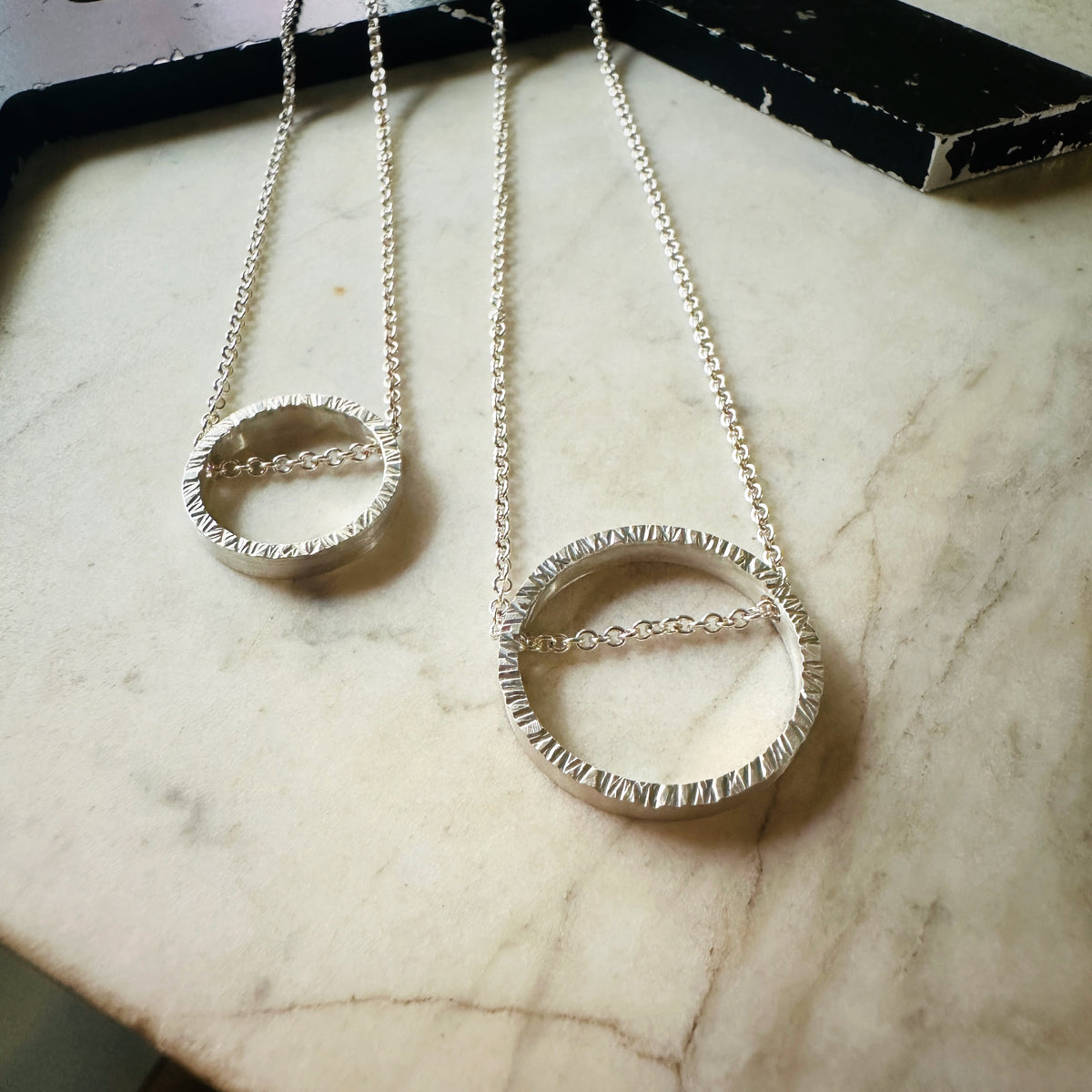 Two hammered silver chain necklaces with textured circular pendant rings resting on a light marble surface.
