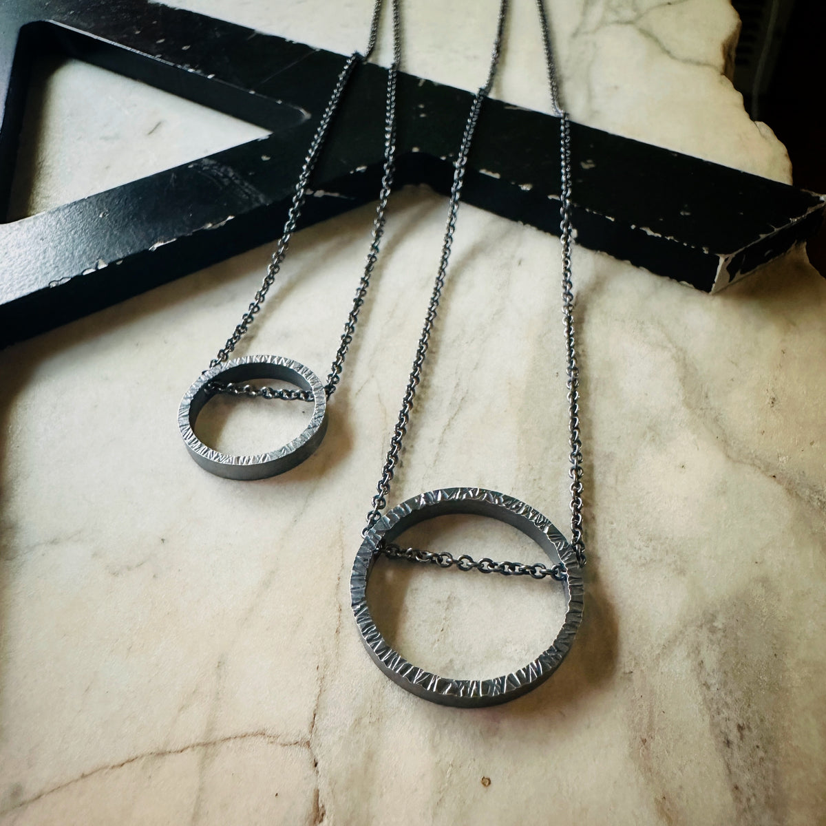 Two oxidized silver textured metal necklaces with circular pendant designs resting on a light beige marble surface next to a black wooden frame.