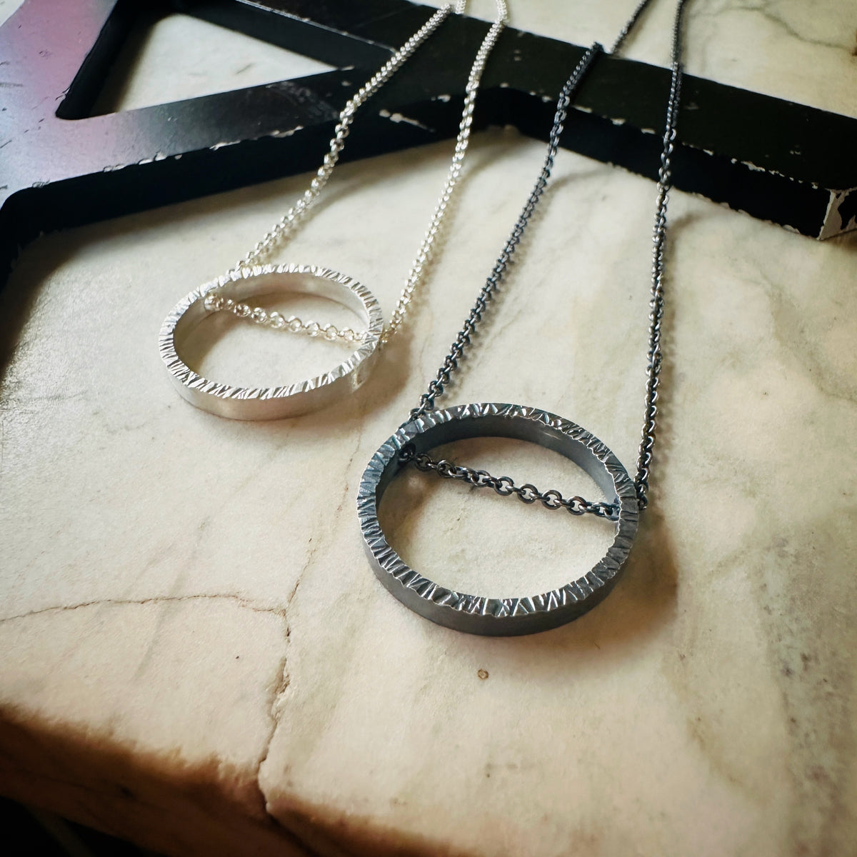 Two minimalist circular pendant necklaces displayed on a light marble surface. One necklace is silver with a textured outer ring and a silver chain, while the other is dark gunmetal gray with a textured outer ring and a dark chain.