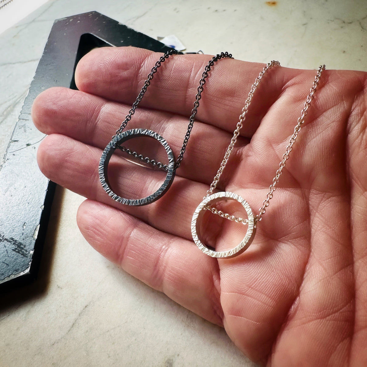 Two textured circular pendant necklaces, one dark silver and bright silver, displayed on a person's hand.