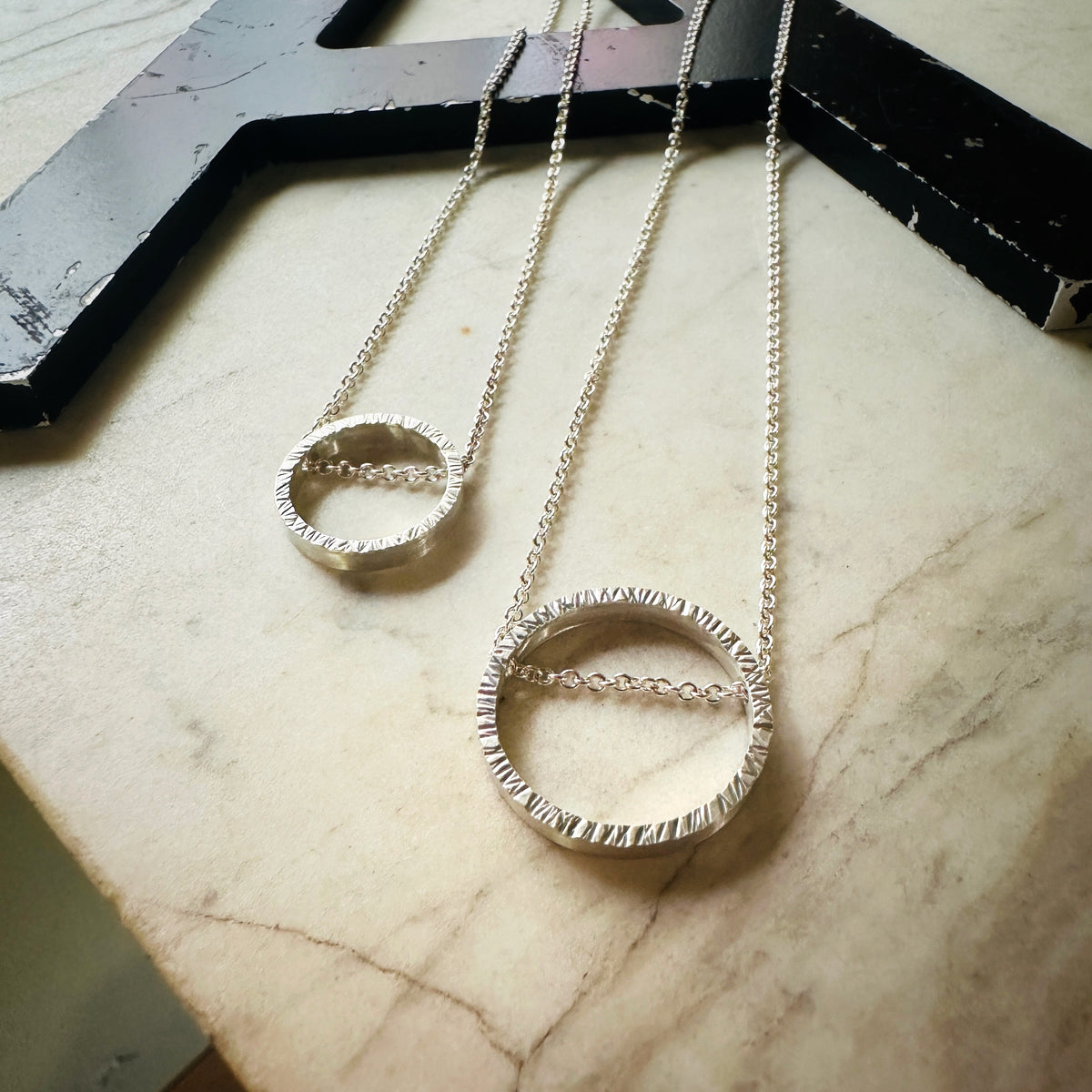 wo hammered silver chain necklaces with textured circular pendant rings resting on a light marble surface.