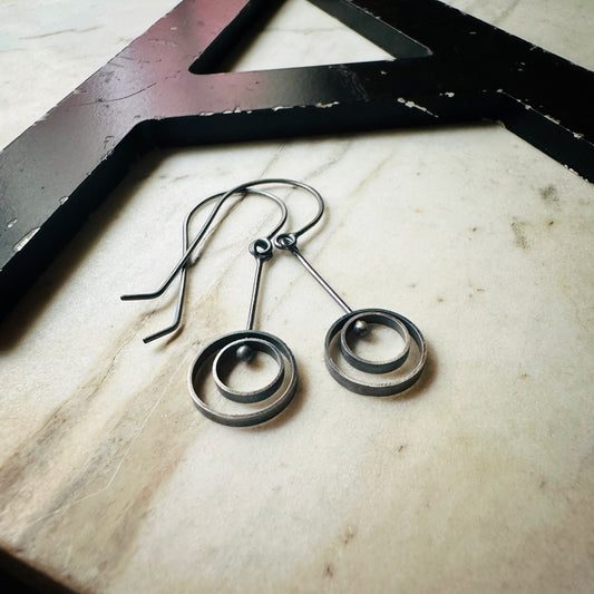 A pair of oxidized sterling silver drop earrings with circular spiral designs, resting on a light beige marble surface next to a black wooden frame.