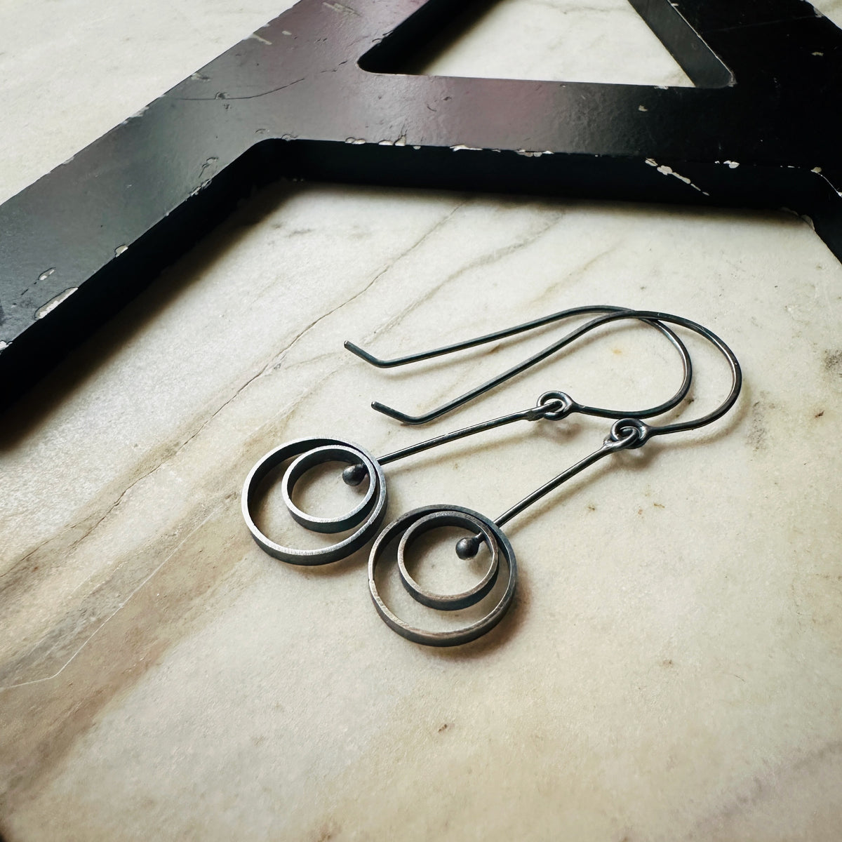 A pair of handmade concentric circle drop earrings made from dark sterling silver resting on a light beige marble surface next to a black decorative letter.