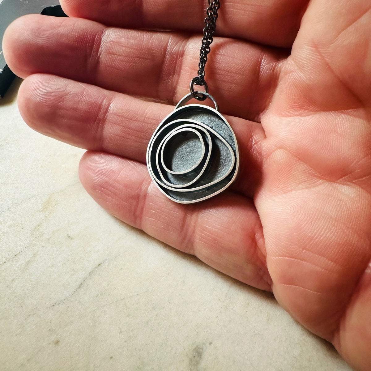 An oxidized sterling silver concentric circle pendant necklace on an oxidized silver chain, resting on a hand for scale