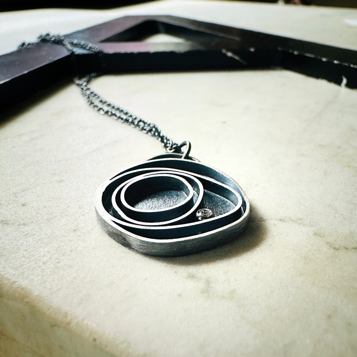 Concentric circle pendant in oxidized silver with a bezel set diamond on a marble base. 