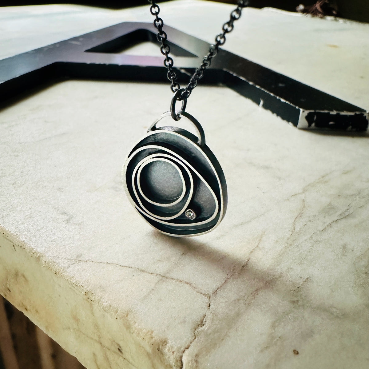 Concentric circle pendant in oxidized silver with a bezel set diamond on a marble base. 