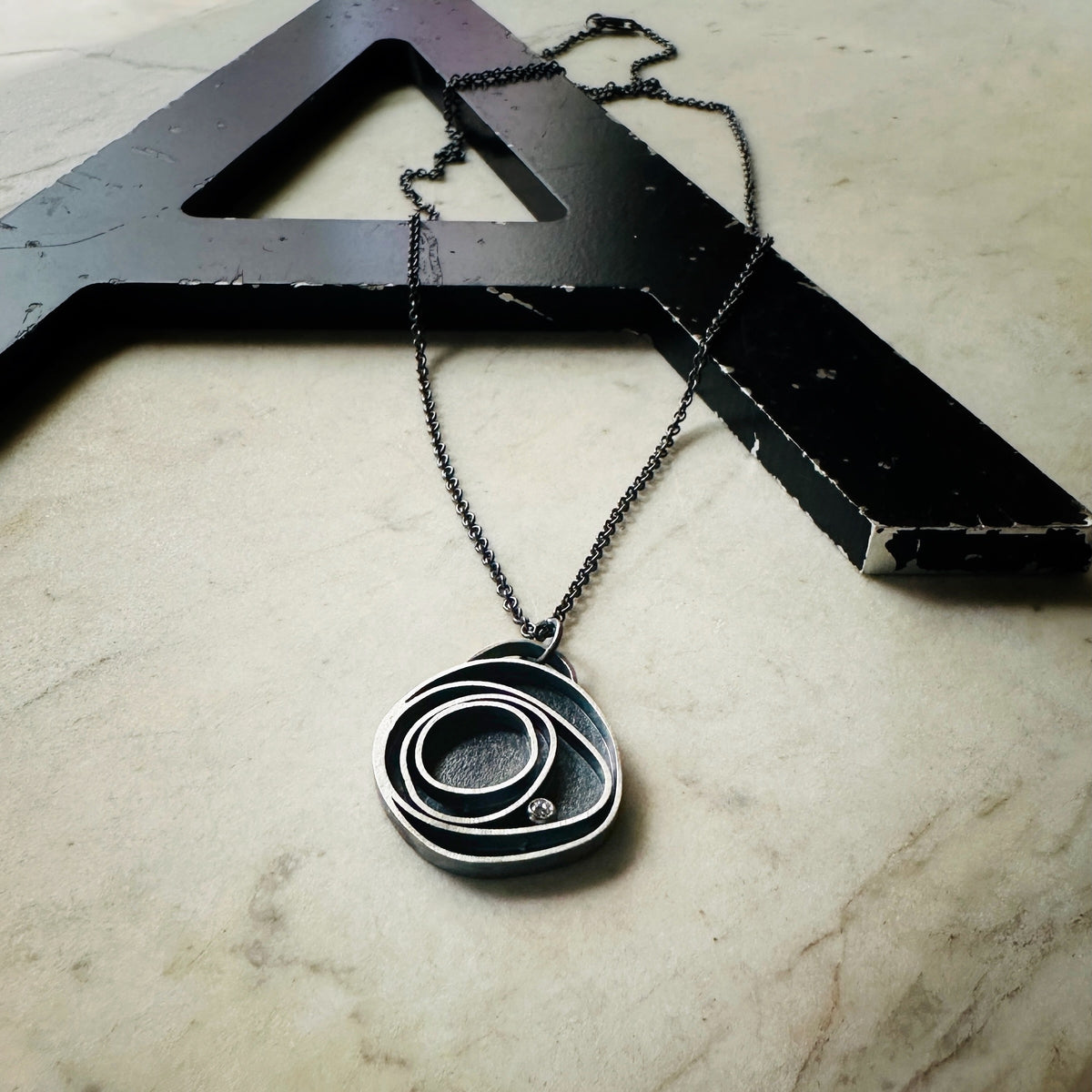 Concentric circle pendant in oxidized silver with a bezel set diamond on a marble base. 