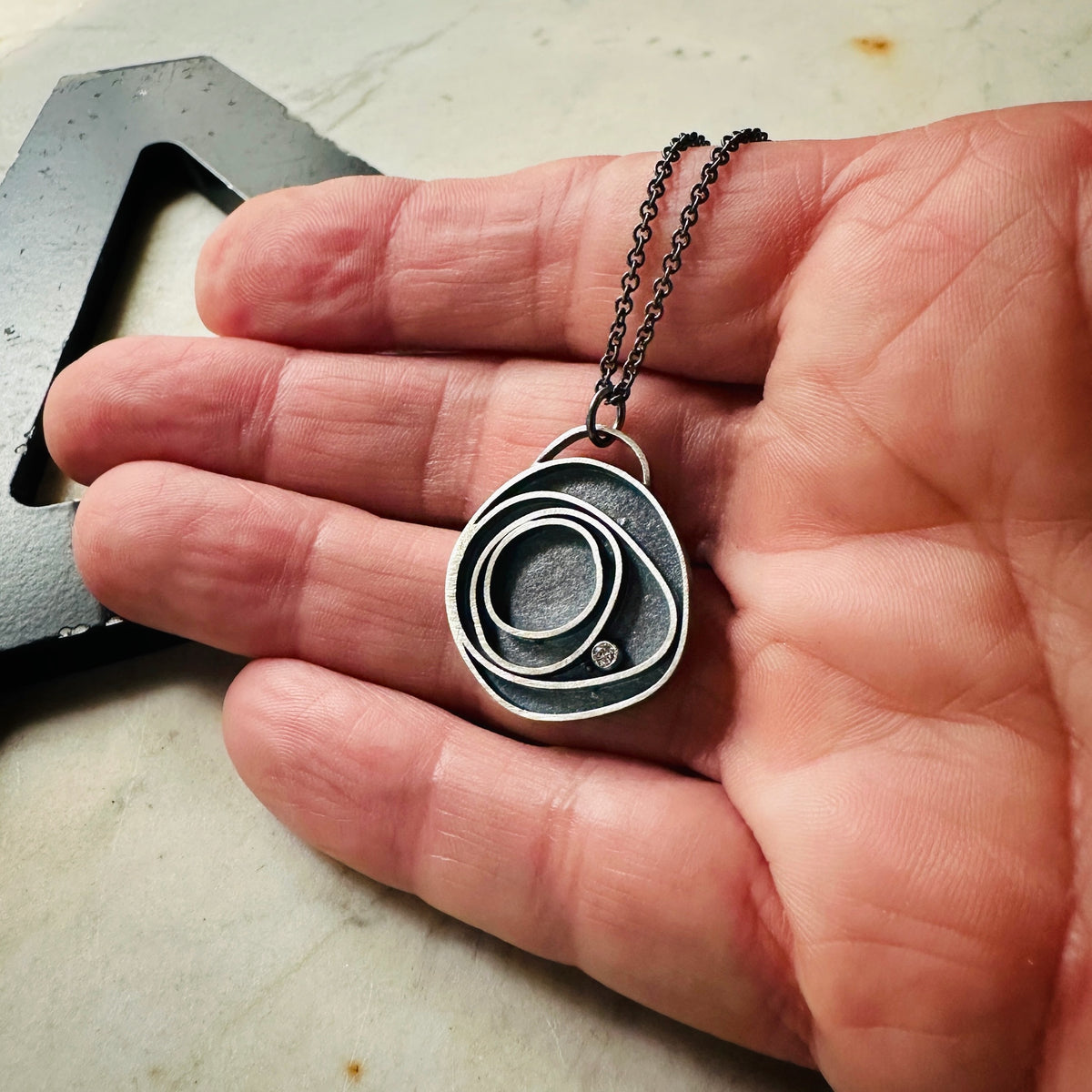 Concentric circle pendant in oxidized silver with a bezel set diamond on a hand. 