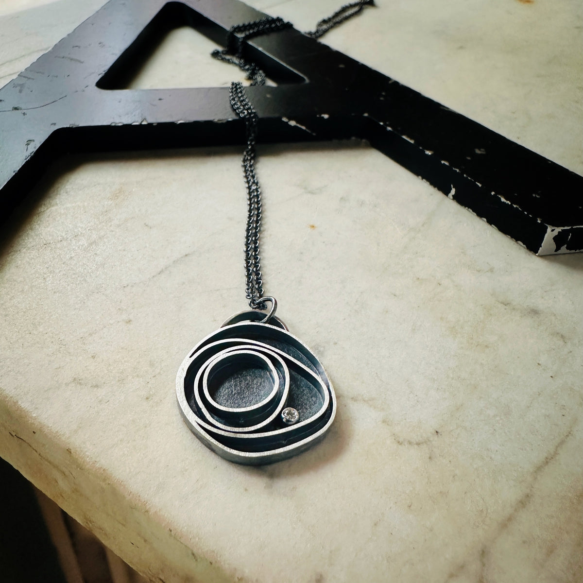 Concentric circle pendant in oxidized silver with a bezel set diamond on a marble base. 