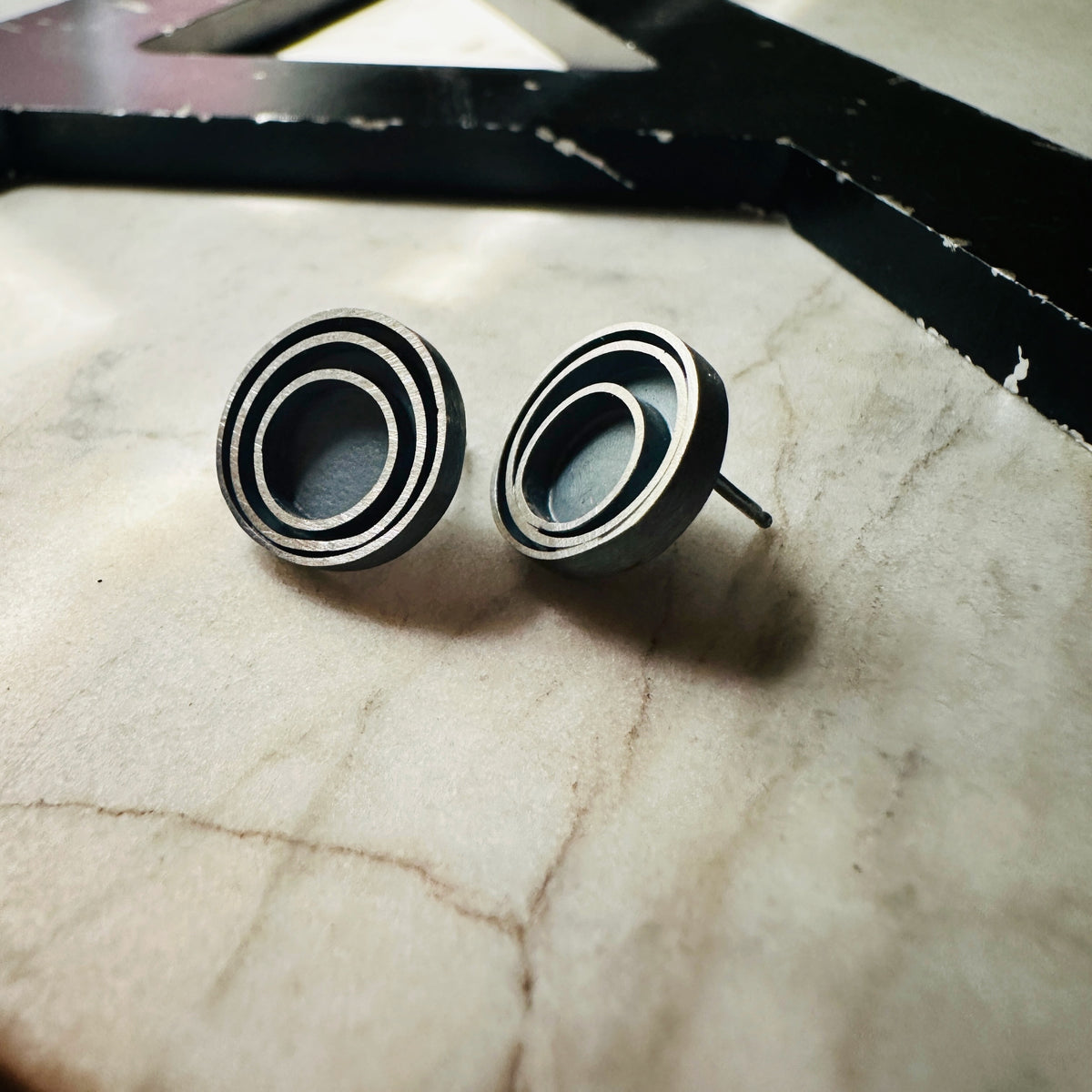 A pair of round stud earrings with a concentric circular design in a oxidized sterling silver, resting on a light beige marble surface.