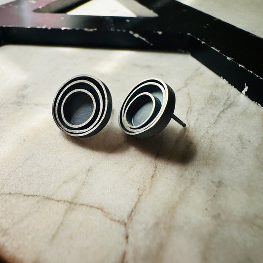 A pair of round stud earrings with a concentric circular design in a oxidized sterling silver, resting on a light beige marble surface.