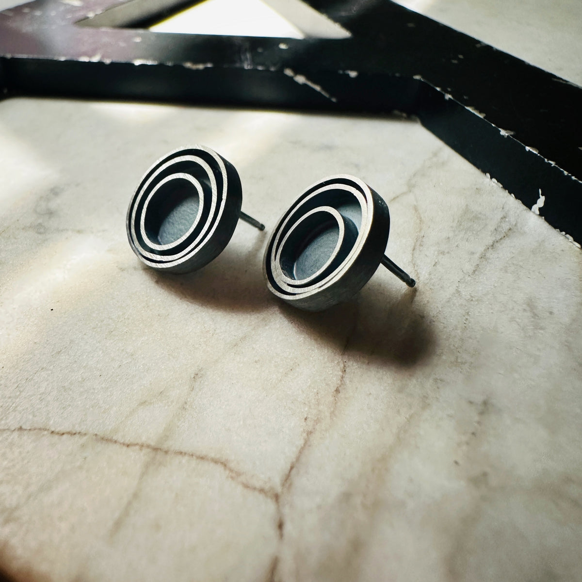 A pair of round stud earrings with a concentric circular design in a oxidized sterling silver, resting on a light beige marble surface.