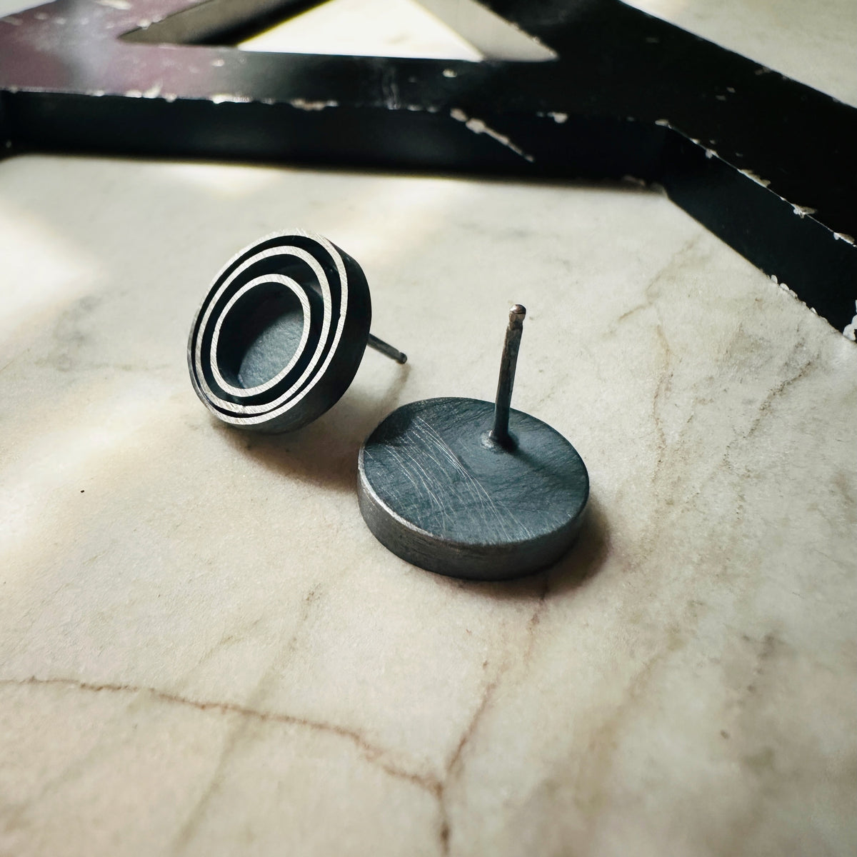 A pair of round stud earrings with a concentric circular design in an oxidized sterling silver, resting on a light beige marble surface. one earring upside down showing the earring post