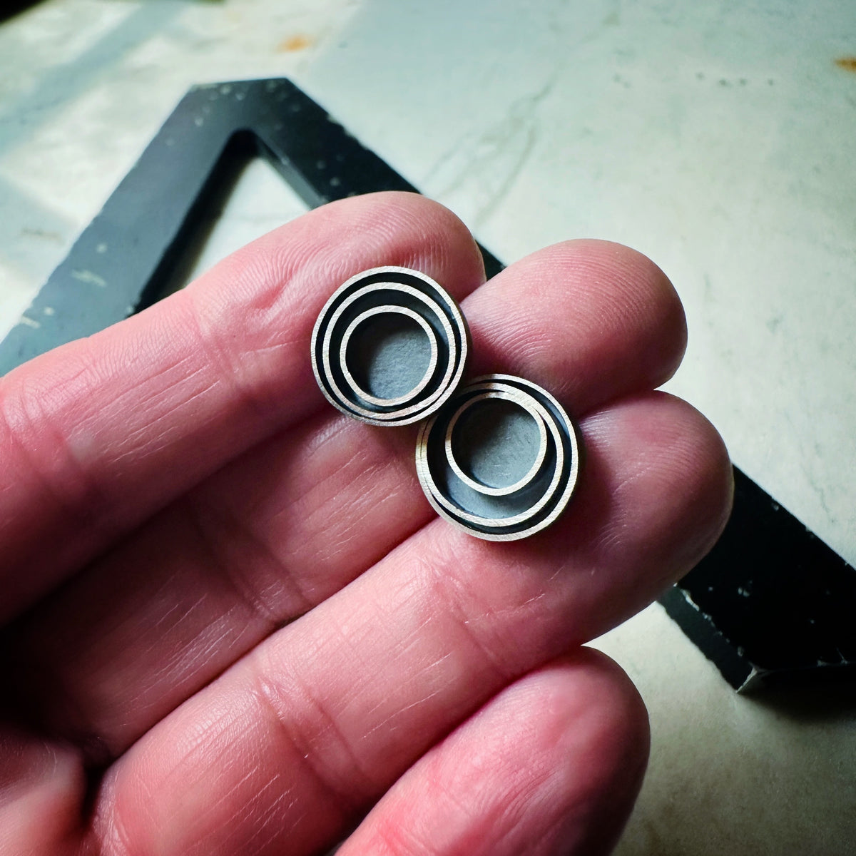 A pair of round stud earrings with a concentric circular design in a oxidized sterling silver, resting on a light beige marble surface held in a hand for scale