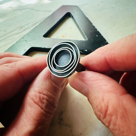 A close-up of a hand holding an oxidized silver ring with concentric circle designs, with a large black metal letter 'A' in the background.