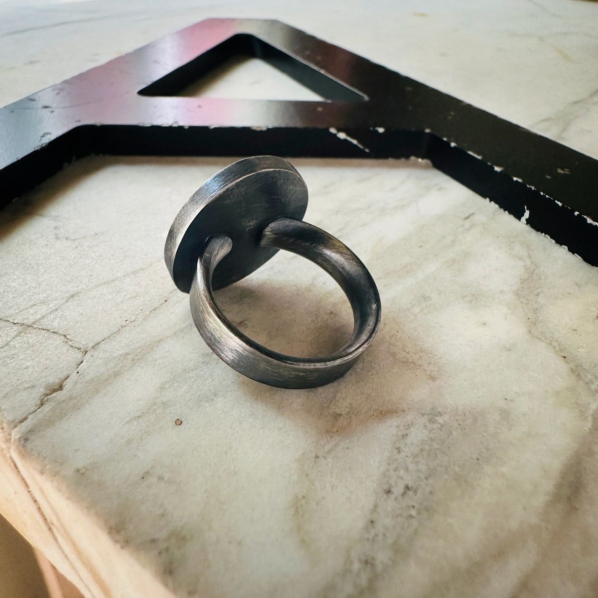 the back of an oxidized silver ring with concentric circle designs, with a large black metal letter 'A' in the background.