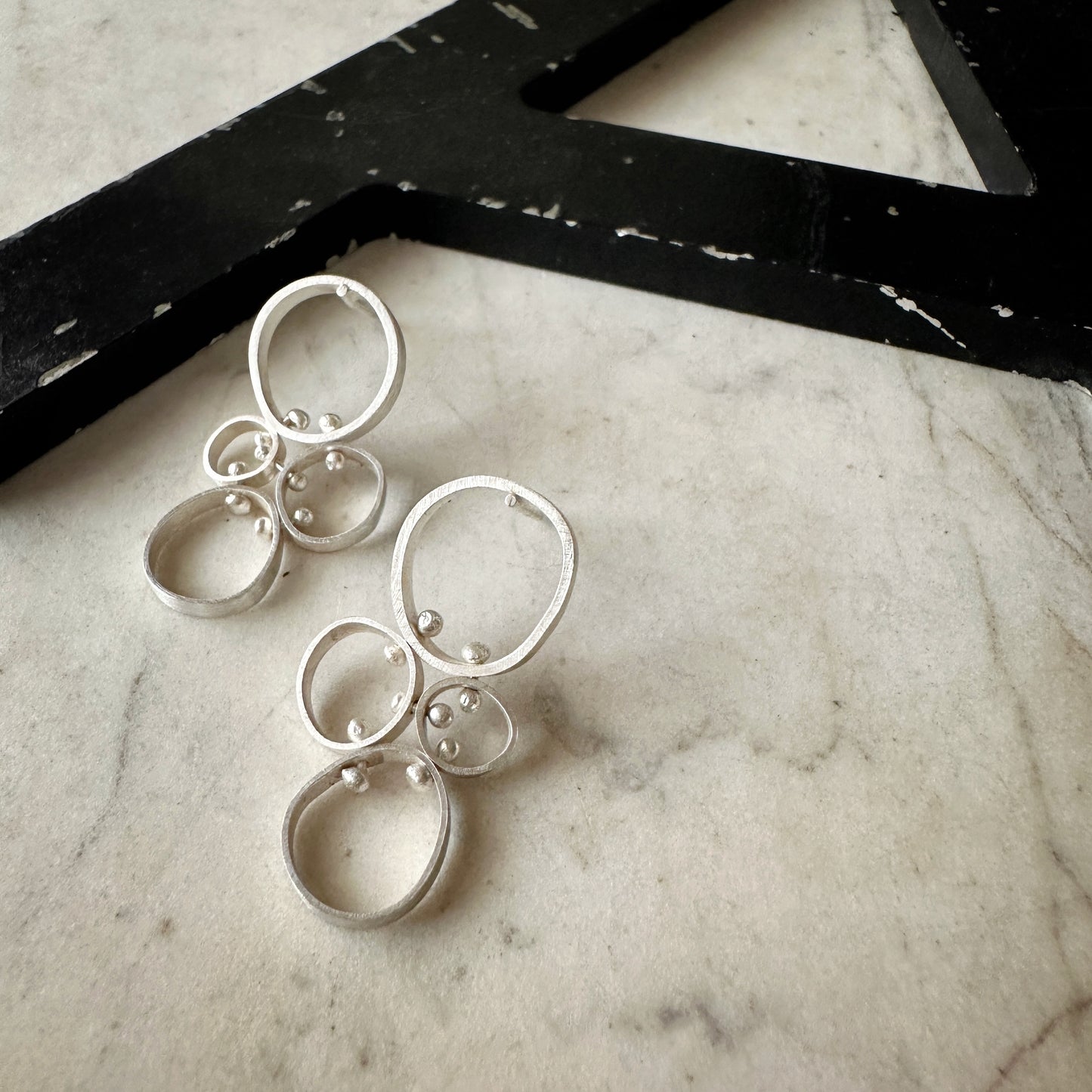 A pair of silver dangle earrings with multiple nested circular rings and small bead details, displayed on a light marble surface next to a black decorative object.