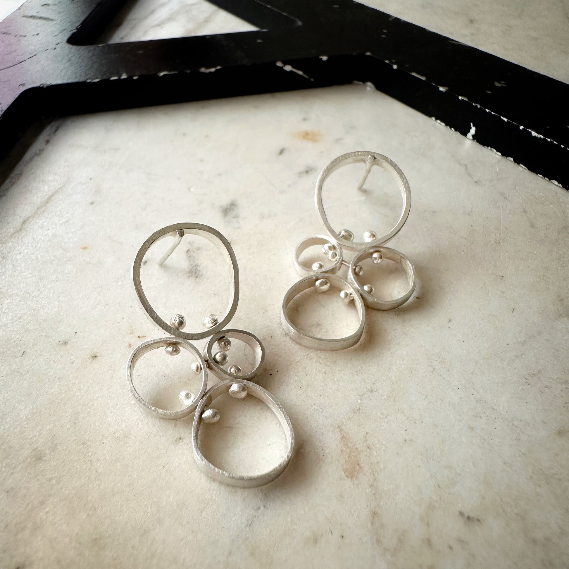A pair of silver-toned geometric dangle earrings with overlapping circular rings and small bead details, displayed on a light marble surface.