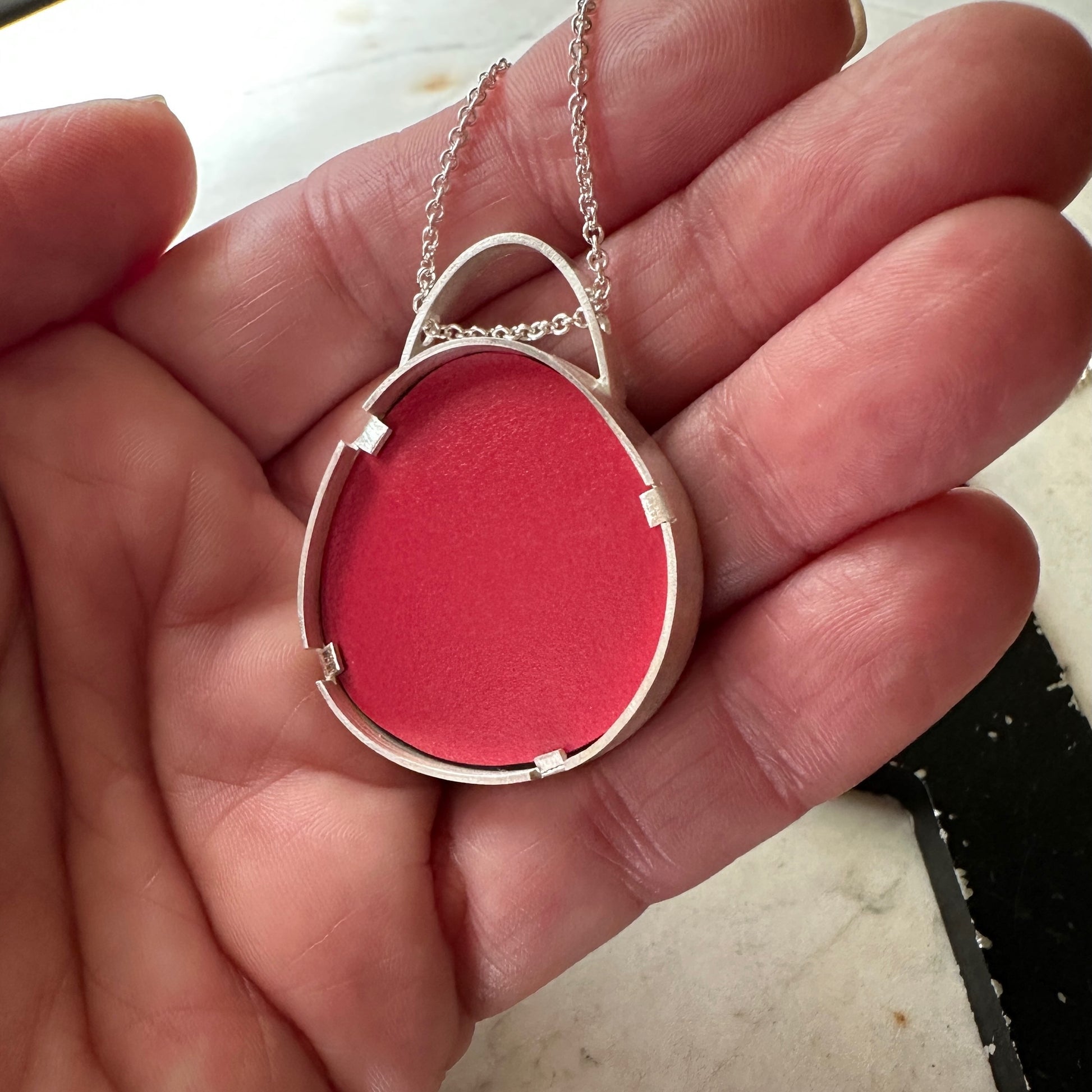 A sterling silver oval pendant necklace with a textured  Formica bright red center, held in a person's hand.