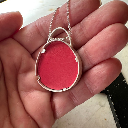 A sterling silver oval pendant necklace with a textured  Formica bright red center, held in a person's hand.