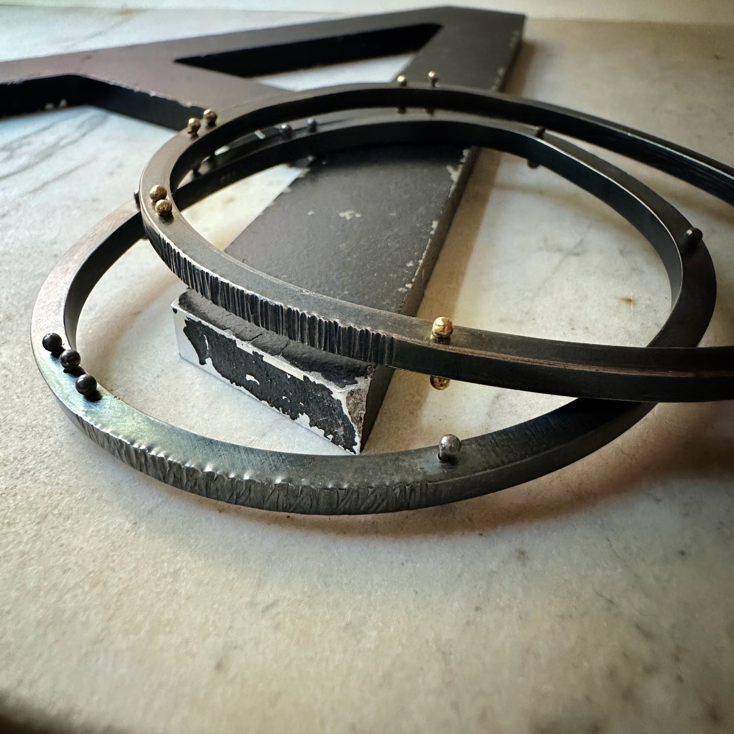 A close-up of a hammer textured oxidized silver bangle bracelet with , resting on a light stone surface. The object features gold and silver small rivets.