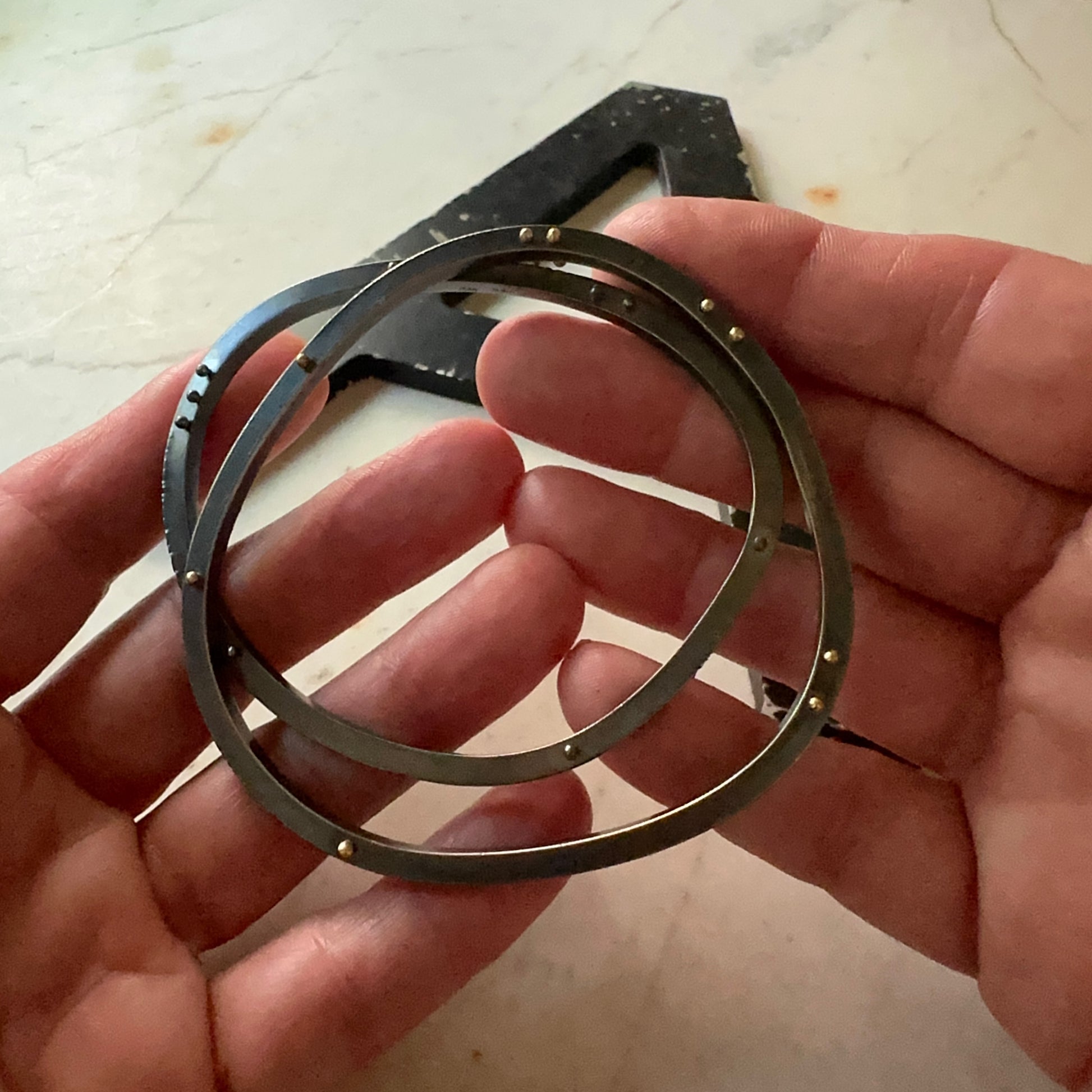 A pair of oxidized sterling silver bangle bracelets held in a person's hands, with a black rectangle visible in the background on a light stone surface.