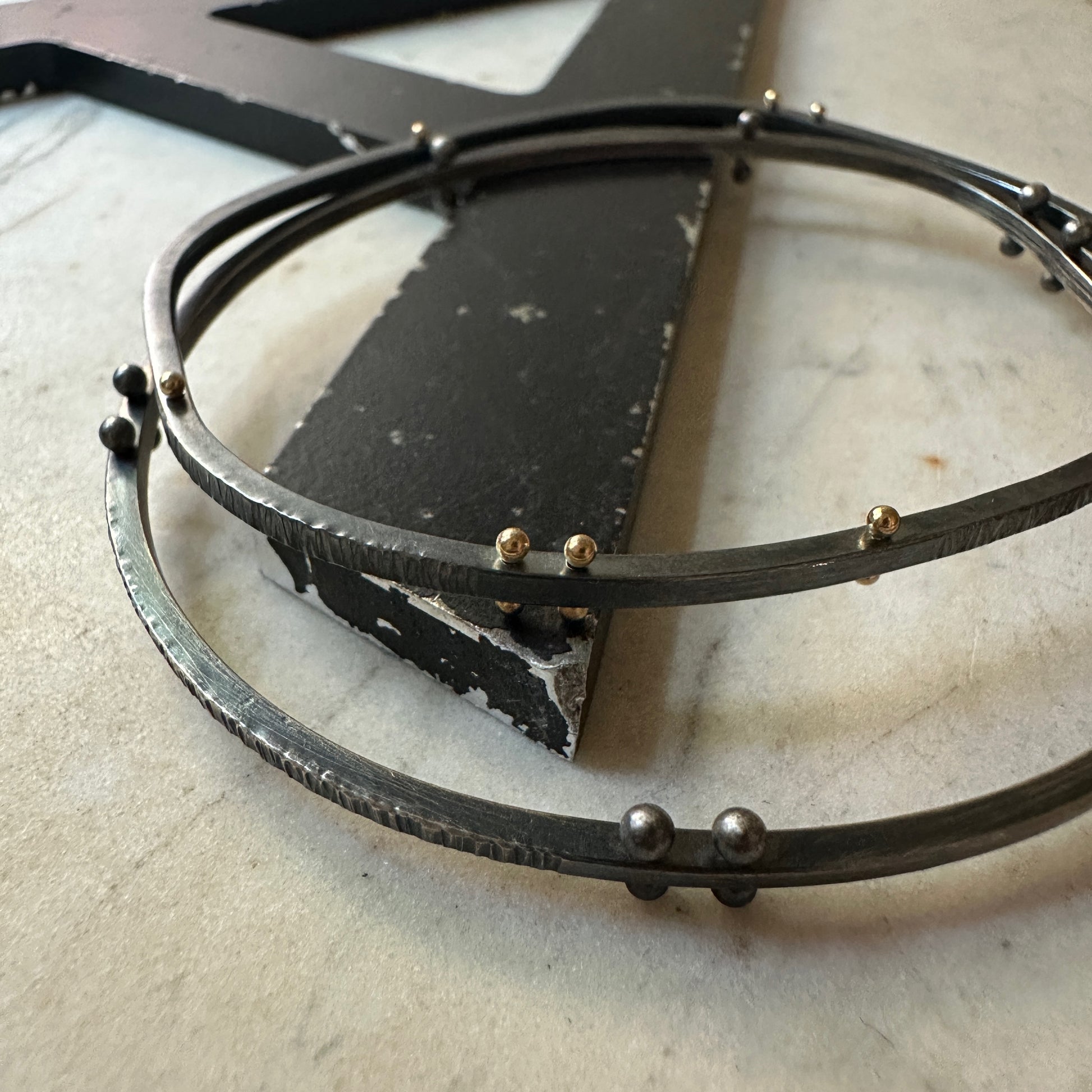 A close-up of oxidized silver bangle with small gold and silver rivet details, resting on a light stone surface next to a black rectangular block.
