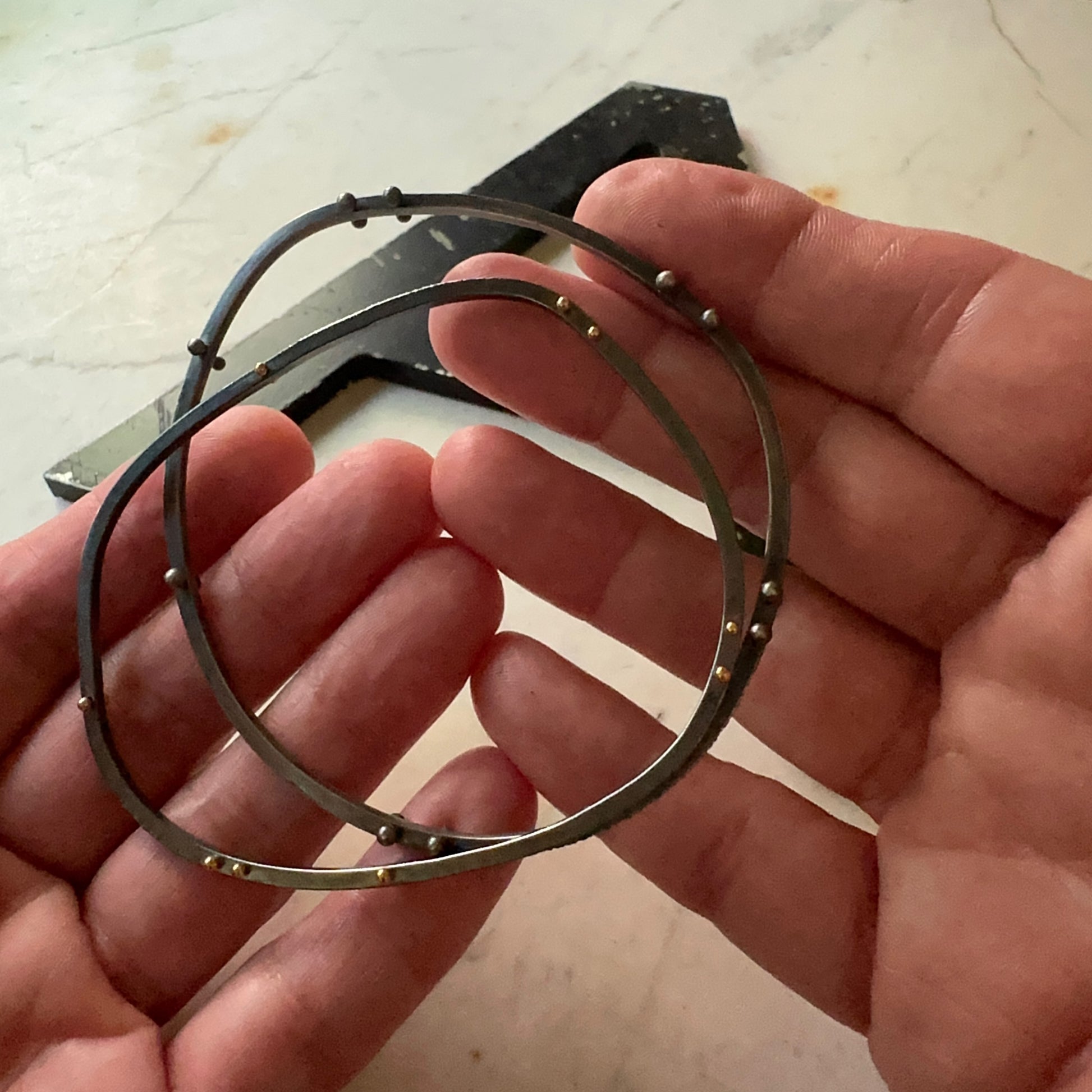 A pair of oxidized silver bangle bracelets with small silver and gold rivet accents, held in a person's hand over a light stone surface.